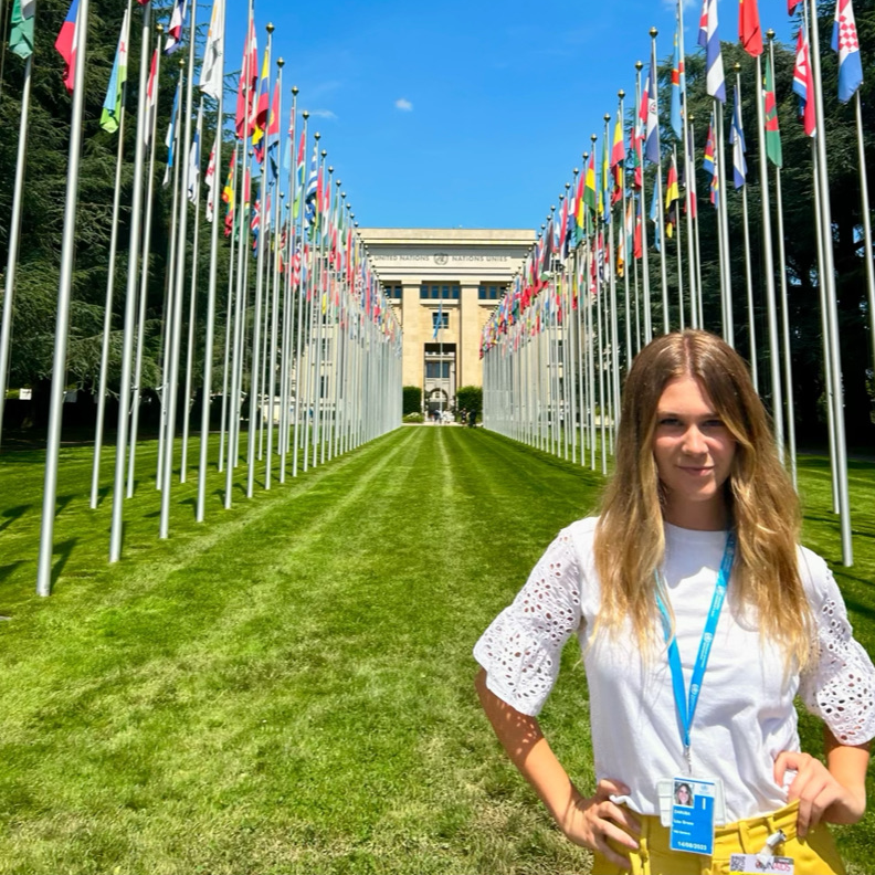 Liza Zaruba, a woman with a light skin tone and long blonde hair, stands in between country flags outside of the United Nations. She rests her hands on her hips and is wearing a white blouse and yellow pants.