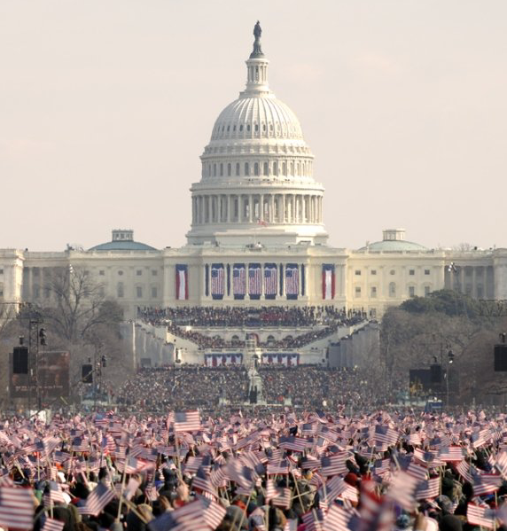A long-distance view of the U.S. Capitol building, draped in U.S. flags. A large crowd in front waves smaller flags in the air. 