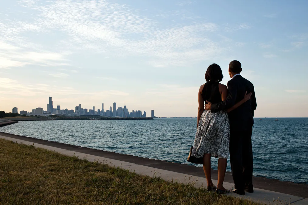 President and Mrs. Obama face Lake Michigan as they look at the Chicago skyline. Their backs are to the camera as they embrace. 