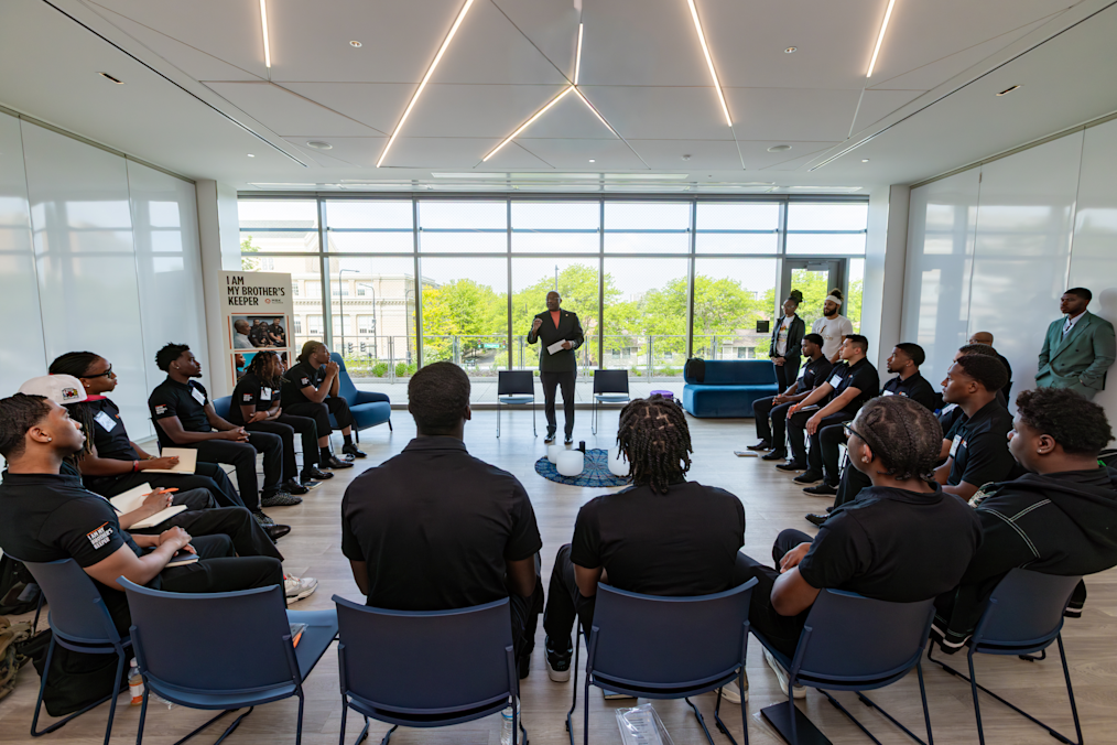 A Black man in a dark jacket speaks to a group of young Black men sitting in a large circle of chairs in a room with floor-to-ceiling windows. A sign on the left reads "I AM MY BROTHER'S KEEPER."