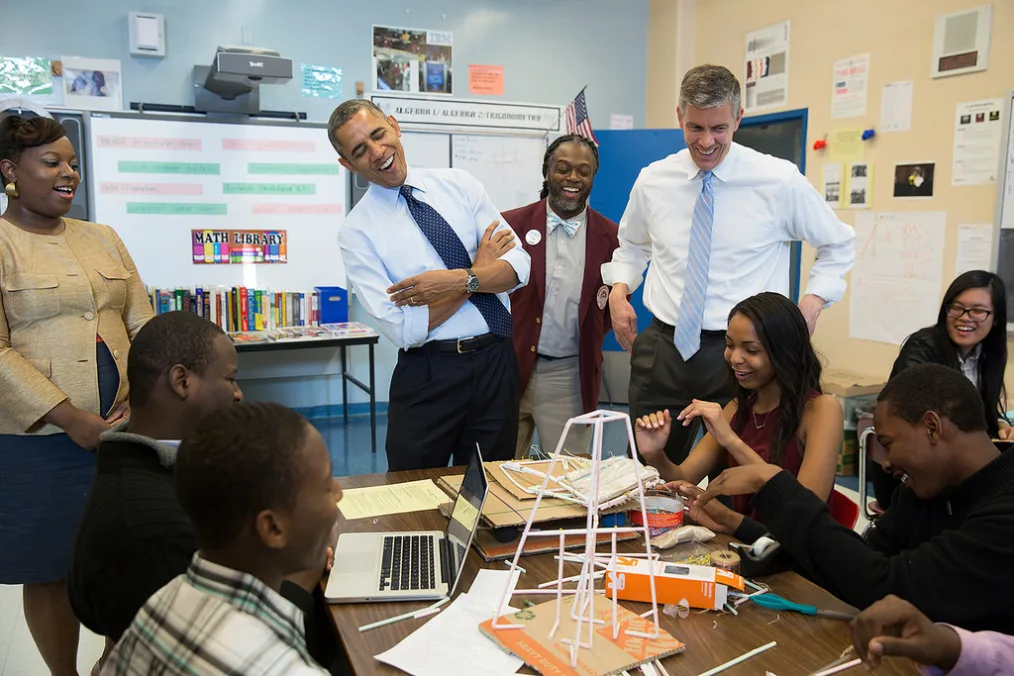 President Obama wearing a light blue button-up shirt, a dark blue tie, and black pants stands with his arms folded and laughs in a room with other individuals of a variety of skin tones. There is a wooden table with a laptop, cardboard, straws, and other craft materials on top. In the background are a white-board, books, and other posters on top of a blue and yellow wall in what appears to be a school classroom. 