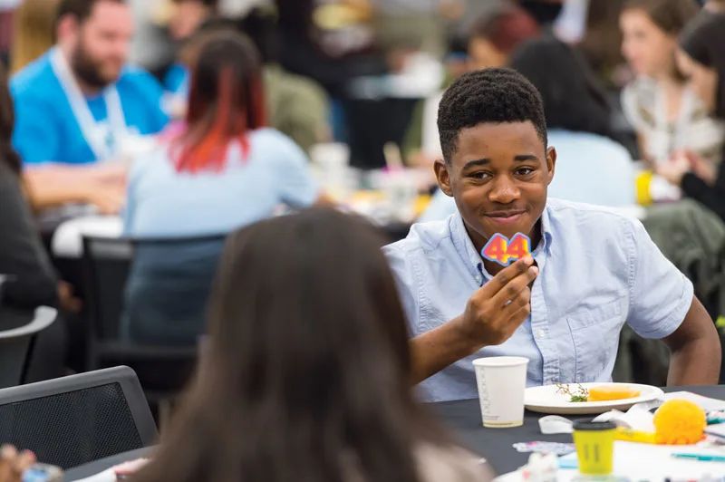 A young man smiles at a peer holding up a sticker that reads 44 at an Obama Foundation Training Day in 2017.