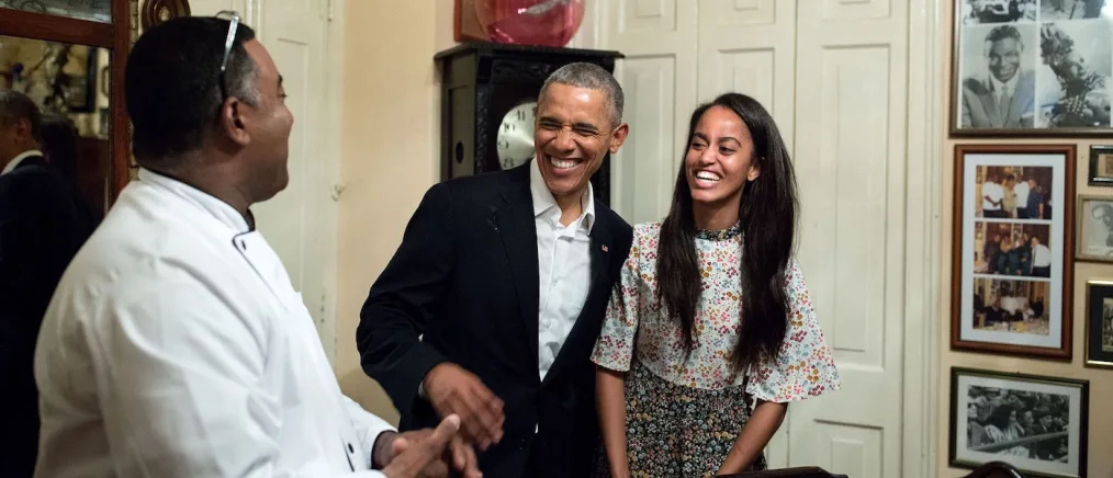 President Obama stands smiling with his daughter Malia talking with a black man with a medium deep skin tone.