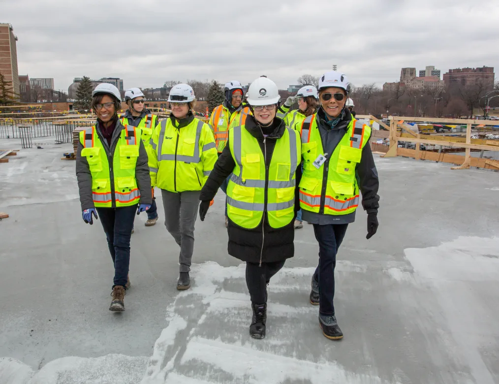 Valerie Jarrett smiles and walks leading a crowd of people with various skin tones at a construction site. All are wearing pants, coats, gloves, and jackets with yellow safety vests and white hard hats. 