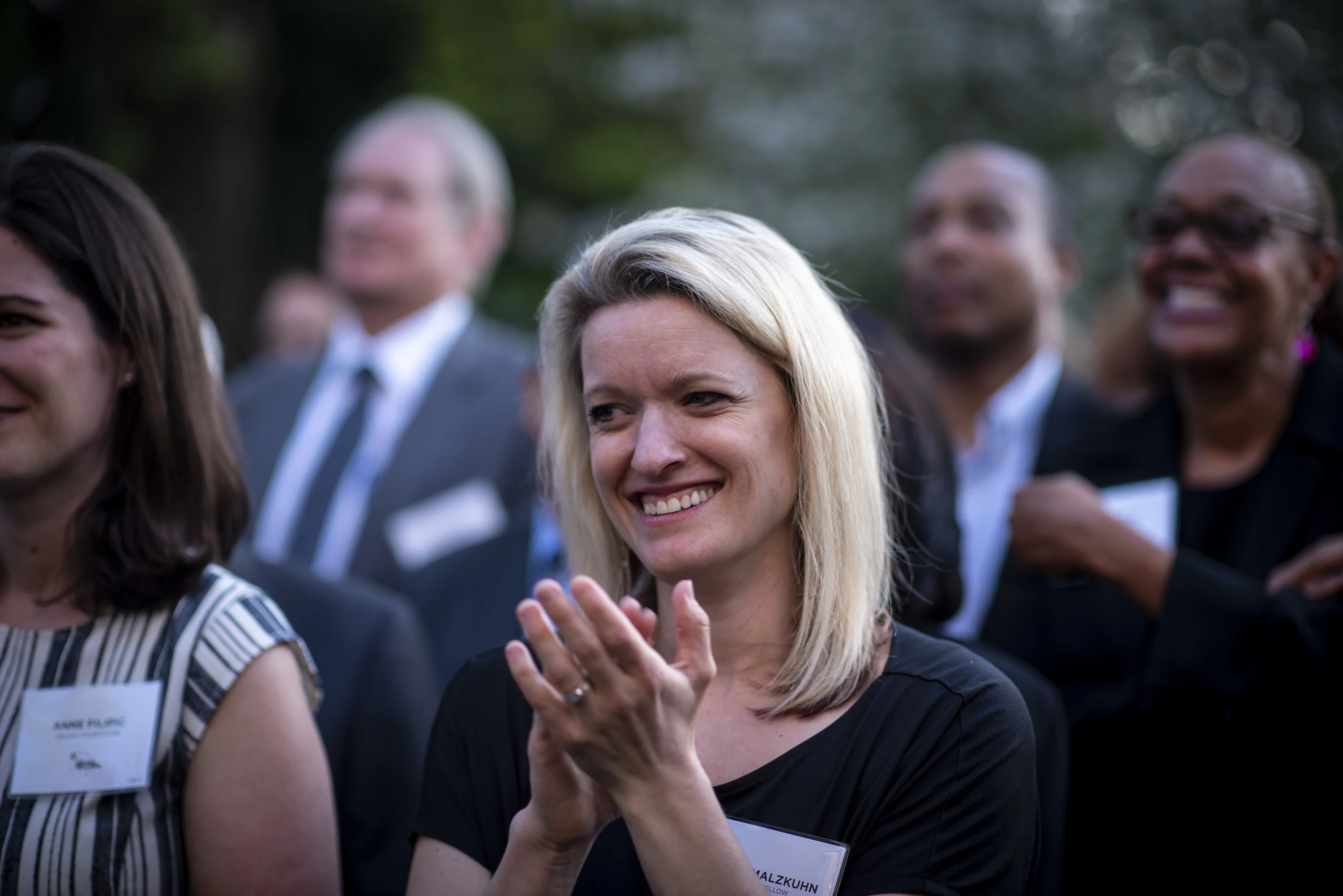 A blonde-haired White woman looks to the left, her hands in a clapping position. People stand nearby but are out of focus. They all wear name tags.