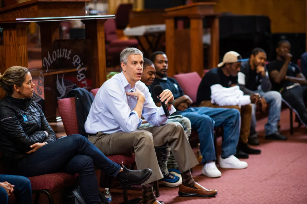 Arne Duncan, former Secretary of Education, holding a microphone addresses a crowd of young men of color.