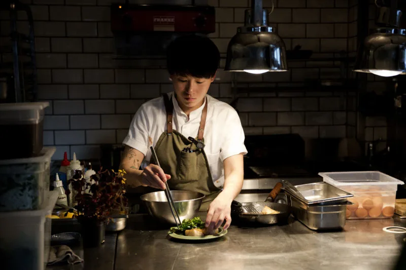 Light skin toned person with ear length black hair standing at a countertop in dark kitchen space. The person is wearing a white short sleeve shirt and green apron, using a large pair of tweezers to place greens and light-colored meat on a green plate. There are sauce bottles, skillets and several clear containers with ingredients on the countertop. 