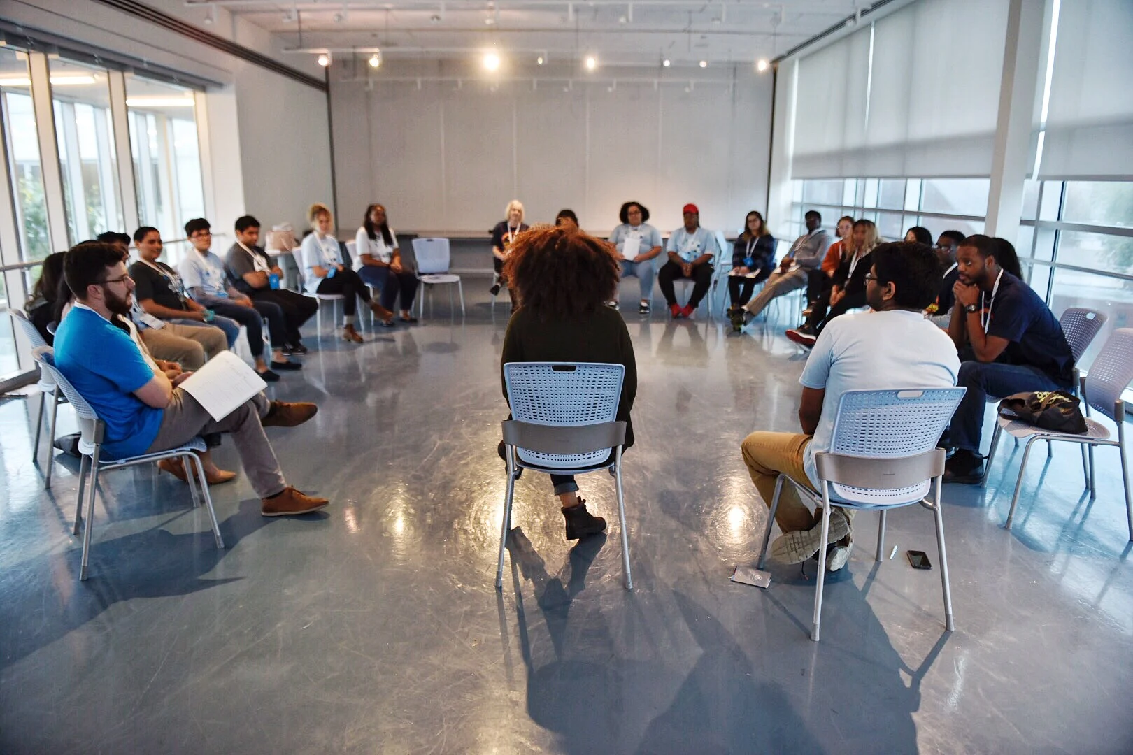 A room full of people with various skin tones sitting in white chairs, in the shape of a circle 