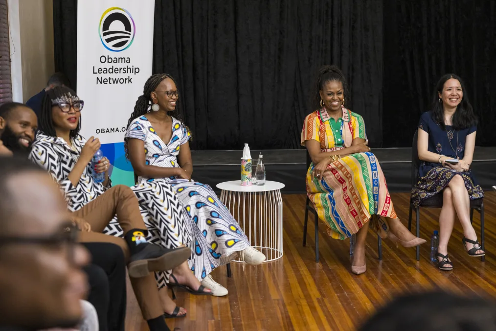 Michelle Obama sits in a circle with women with a range of light to deep skin tones and Michael Strautmanis. Mrs. Obama is wearing a multicolored dress and sits with her legs crossed. A sign in the background reads, “Obama Leadership Network.” 