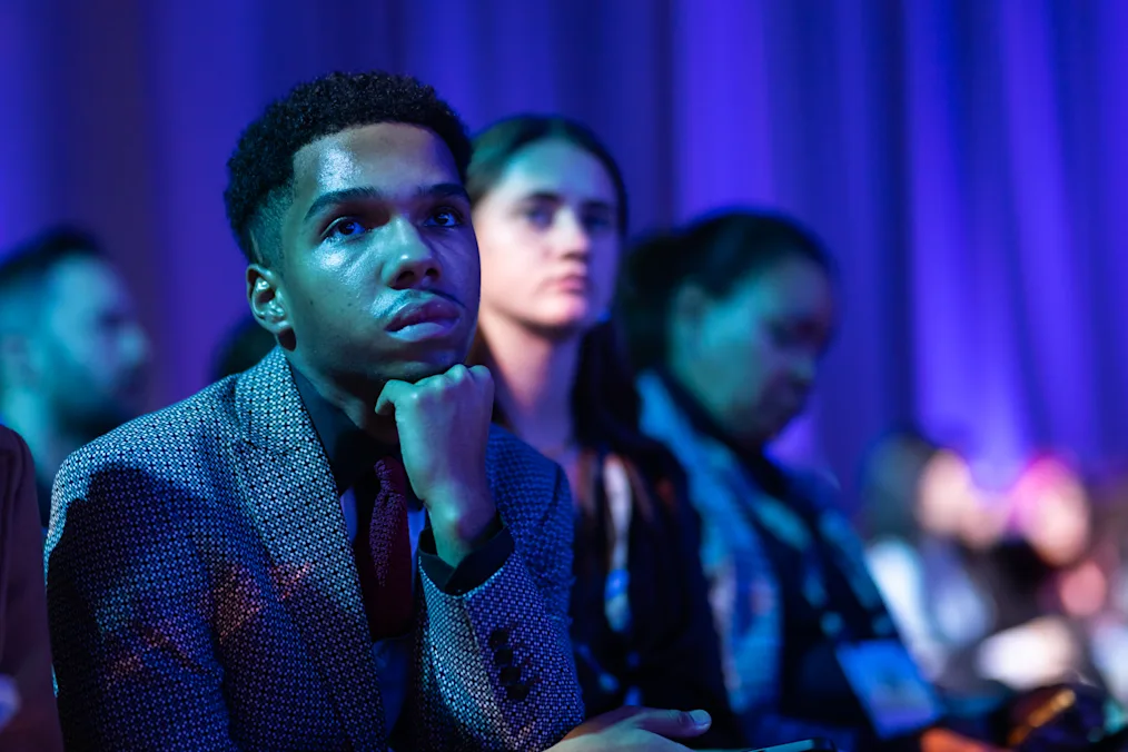 A man with a medium skin tone rests his fist on his chin as he listens from an audience. He is sitting next to a man and two women with light to medium skin tones. They are in a dimly lit blue room.