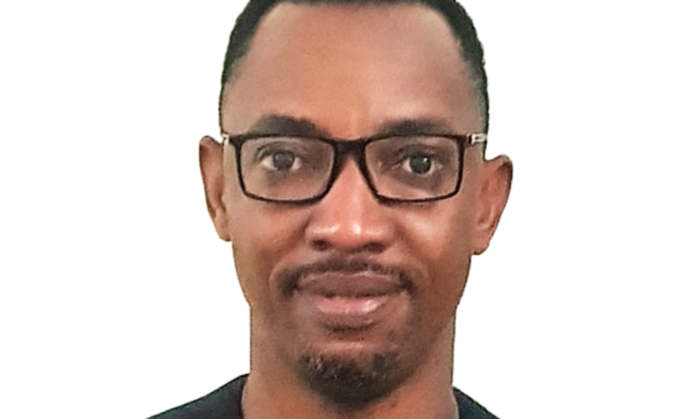 Edwin Ikhuoria, a black man with a medium-deep skin tone, low cut black hair, beard, and glasses wearing a black shirt, looks into the camera.