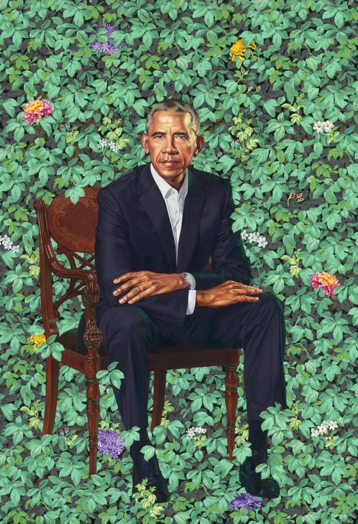 The White House portrait of President Obama sitting down in front of a background with flowers. He is wearing an all black suit with a white button up shirt.