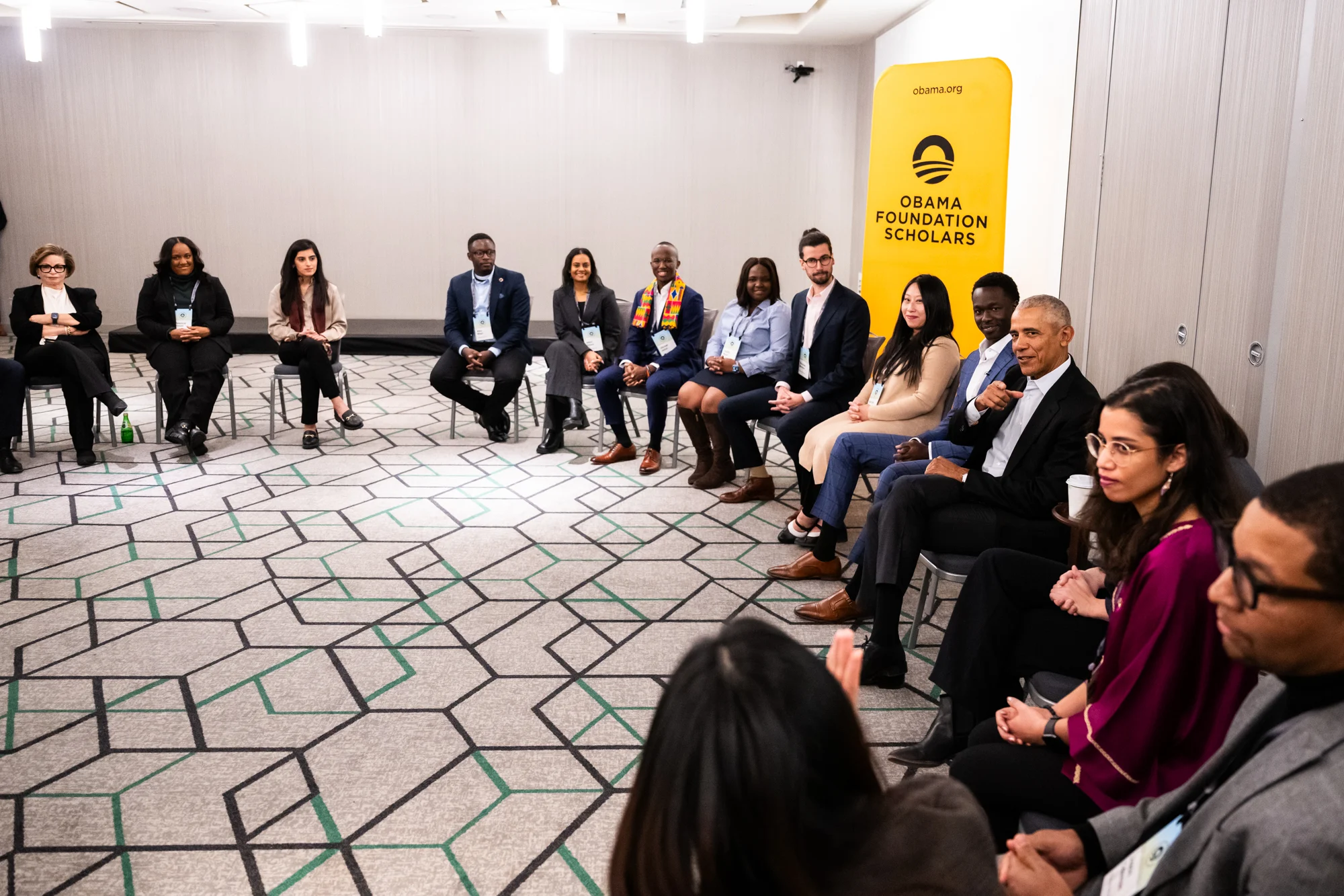 Obama Scholars with a range of light to dark skin tones sit in a semi circle with Valerie Jarrett and President Obama. All are dressed professionally. A sign in the background reads, “Obama Foundation Scholars.”