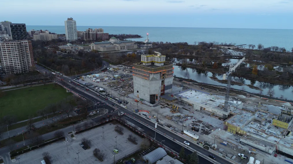 An aerial view of the site of the Obama Presidential Center on a foggy day. A concrete structure with scaffolding is in the middle with the Museum of Science and Industry, lagoon, Jackson Park, and Lake Michigan in the background, and the east end of Midway Plaisance to the left.