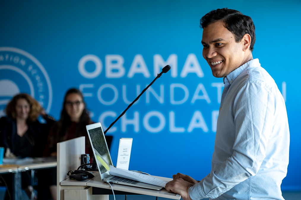 Juan Carlos Monterrey Gómez, a man with a medium skin tone, speaks behind a podium. He is smiling. Two women are blurred in the background. On the side of him, a blue wall reads, “Obama Foundation Scholars.”