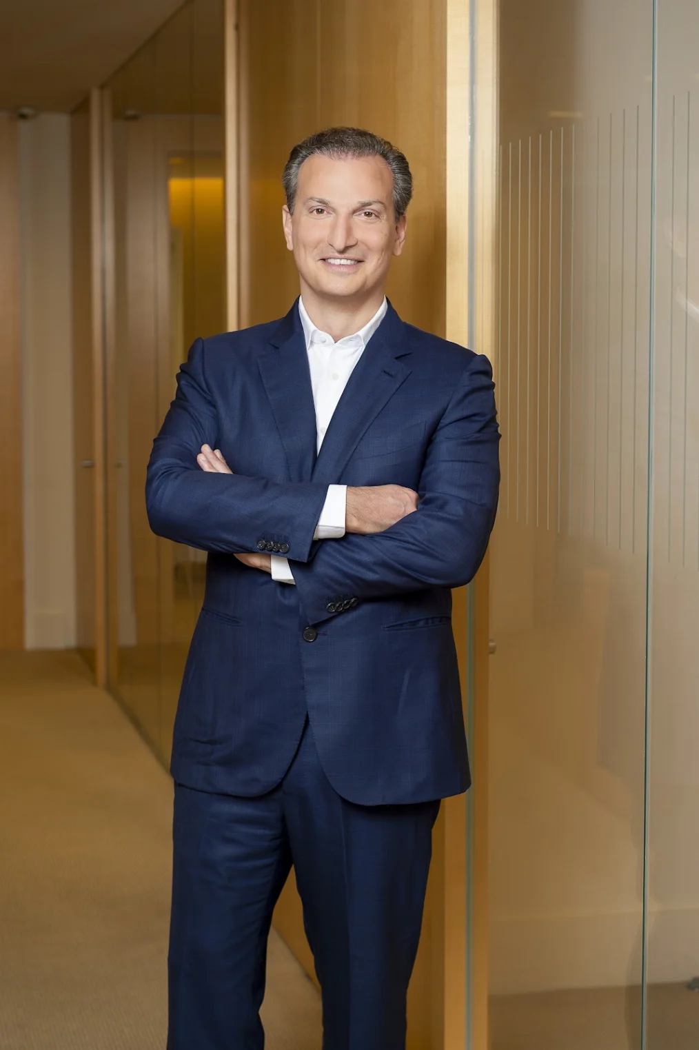 A white man with gray hair wearing a blue suit. He smiles for the camera. His arms are crossed. There is a golden color to the background.