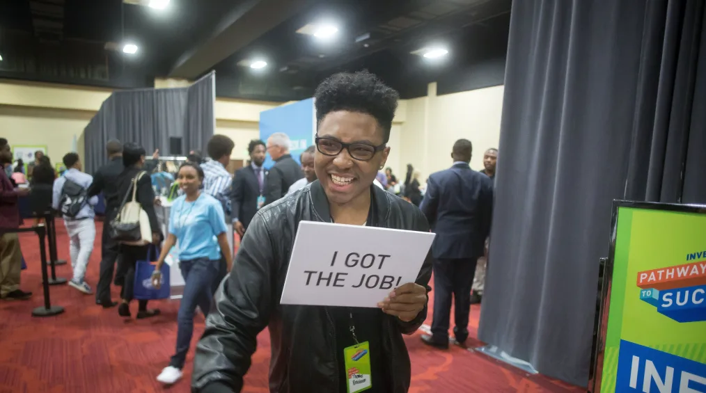 A person with medium-dark skin tone, curly hair and glasses holds a white sign with the words "I GOT THE JOB!" Behind him, people mill around in what appears to be a conference setting with draped panels. A bright green sign in the bottom right with the words "Pathways to Success" partly cut off by the edge of the photo.