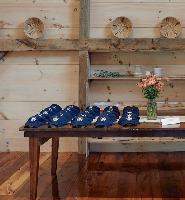 In a rustic wooden room, a group of blue hats line a table next to a vase of flowers. The hats feature a white Obama Foundation rising sun logo. 