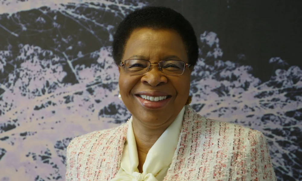 Graça Machel, a black woman with a medium-deep skin tone, low cut hair and thin metal glasses wearing a white and pink tweed jacket and white blouse smiles toward the camera.