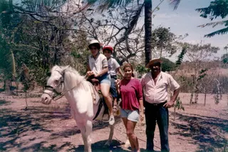 A young Juan Carlos Monterrey Gómez sits on a white horse behind his brother. Beside him are his mom and dad. All have medium skin tones and are smiling.
