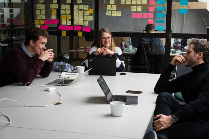 Three people sit around a large white table with laptops and coffee cups. The wall behind them is covered in colorful sticky notes.