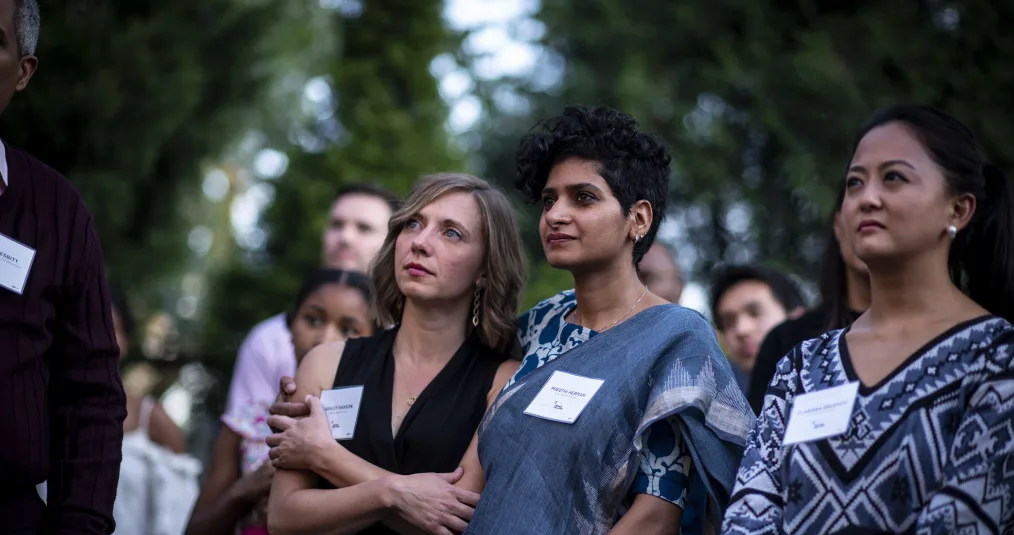 Three women look to the left with a group of people behind them. Two of the women hold each other in a side embrace. All three have name tags identifying them as 2018 Obama Fellows. 