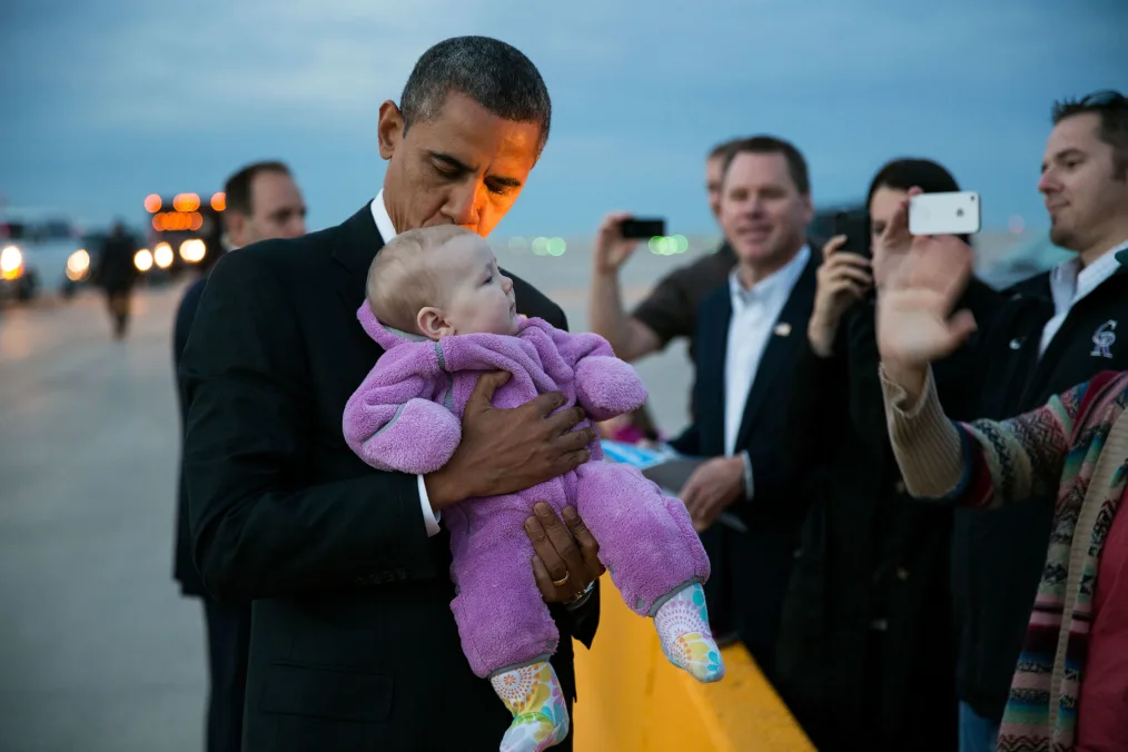 President Obama holds a baby with a light skin tone wearing a purple onesie and colorful boots. The background features people of various skin tones in the background taking photographs on their phones.
