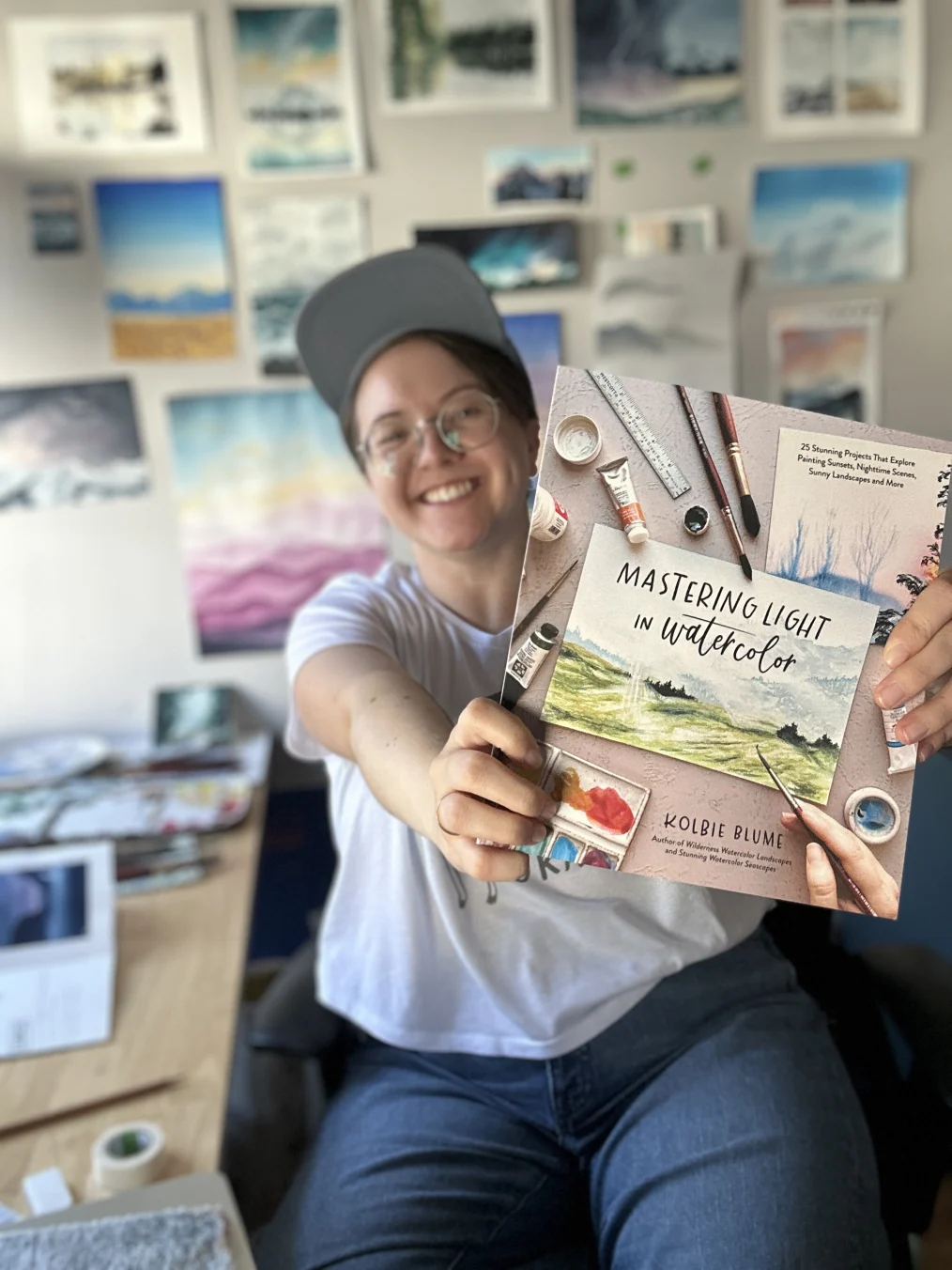 Kolbie Blume is sitting in a chair holding a book and smiling at the camera. Behind them is a light colored wall with more than ten different watercolor paintings handing on the wall. Kolbie has a pale complexion and light colored hair. They are wearing silver round glasses, a grey baseball cap, a white t-shirt, and blue pants. In Kolbie's hand is a book with the words, "Mastering Light in Watercolor, Kolbie Blume." The cover of the book has a canvas and several art tools.