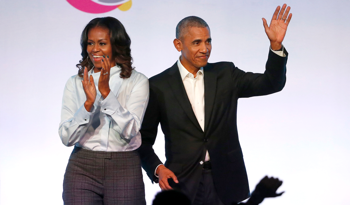 President Barack Obama stands and waves out to a crowd alongside First Lady Michelle Obama as she claps toward the same crowd.