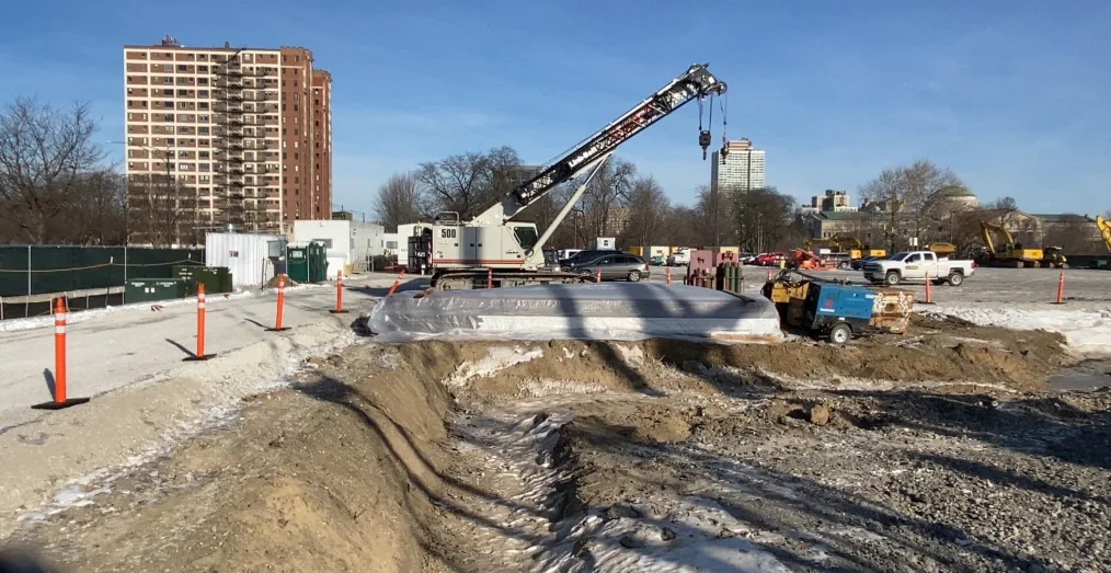 A white crane with a ditch of dirt in the foreground. A building is on the left, a parking area on the right.