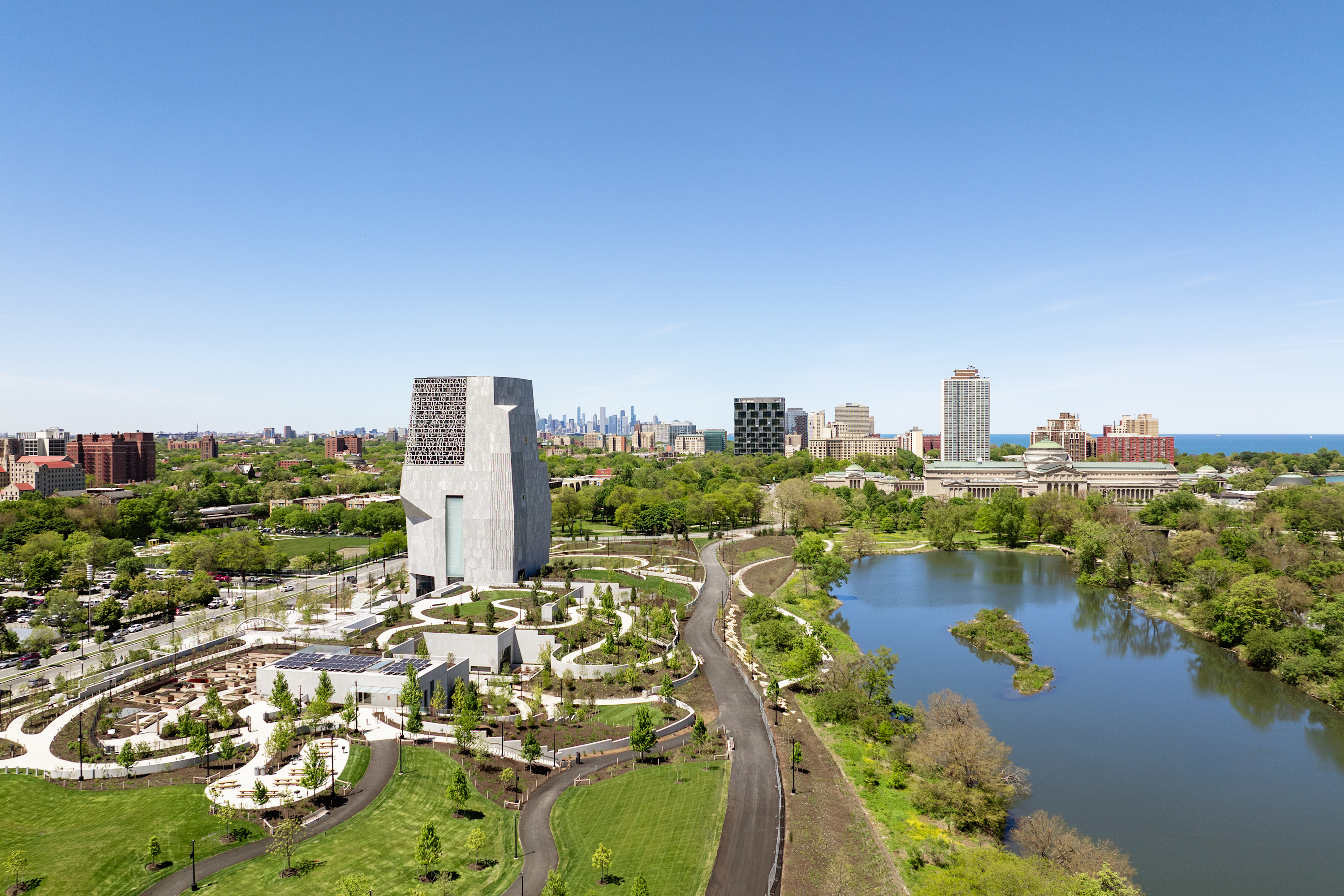 An aerial photo shows the Obama Presidential Center campus. A tall, light gray stone tower rises on the left side near grassy fields and paths. On the right, a calm, blue pond sits next to a large building with a dome. High-rise buildings line the horizon under a clear sky.


