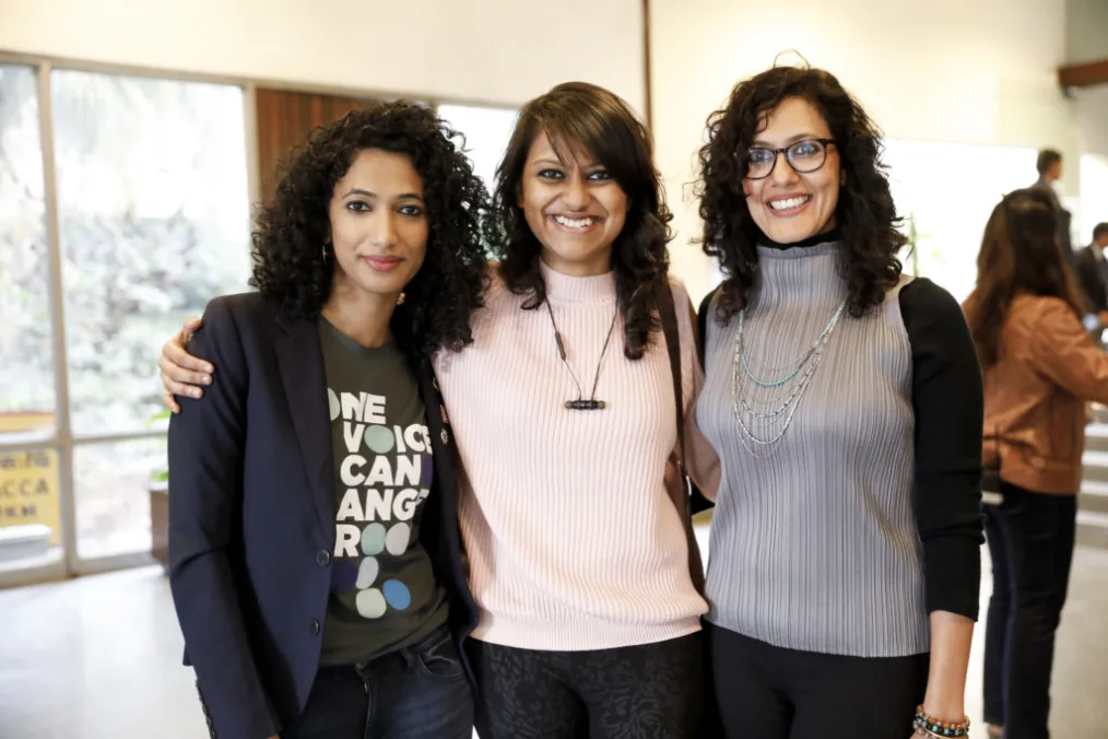 Three women stand side-by-side smiling. They have a range of medium and light skin tones, with shoulder length, and dressed in business casual clothing with a mix of dark and bright colors.