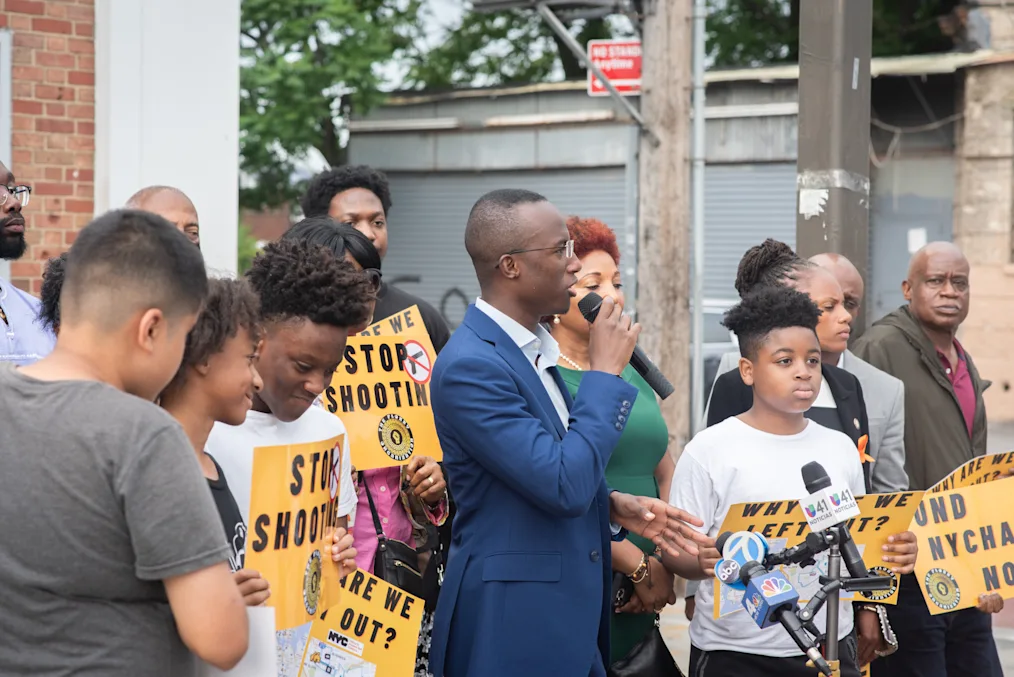Jibreel Jalloh, a Black male with a dark skin tone, holds a microphone as he speaks at a gun violence prevention rally in Canarsie, Brooklyn. He is surrounded by protestors with a range of light to dark skin tones holding posters. Jabreel is wearing a navy suit. 