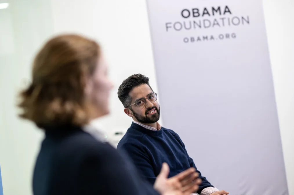 Nathan Blake has a medium skin tone and beard, and smiles at a woman with short hair who is out of focus. He wears a navy sweater over a red and white checked shirt and eyeglasses. “Obama Foundation, Obama.org” is written on a white banner behind him.