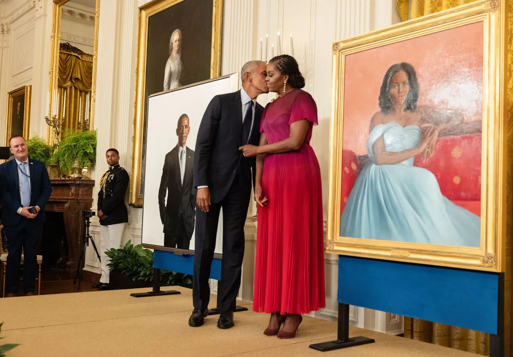 President Barack Obama wearing a black suit with a blue and white vertically striped button-up shirt and blue tie kisses First Lady Michelle Obama, wearing a red dress and brown heels, on the cheek. The are two portrait paintings of each of them on opposite sides of the couple. There are also other paintings and people of different skin tones in the background. 