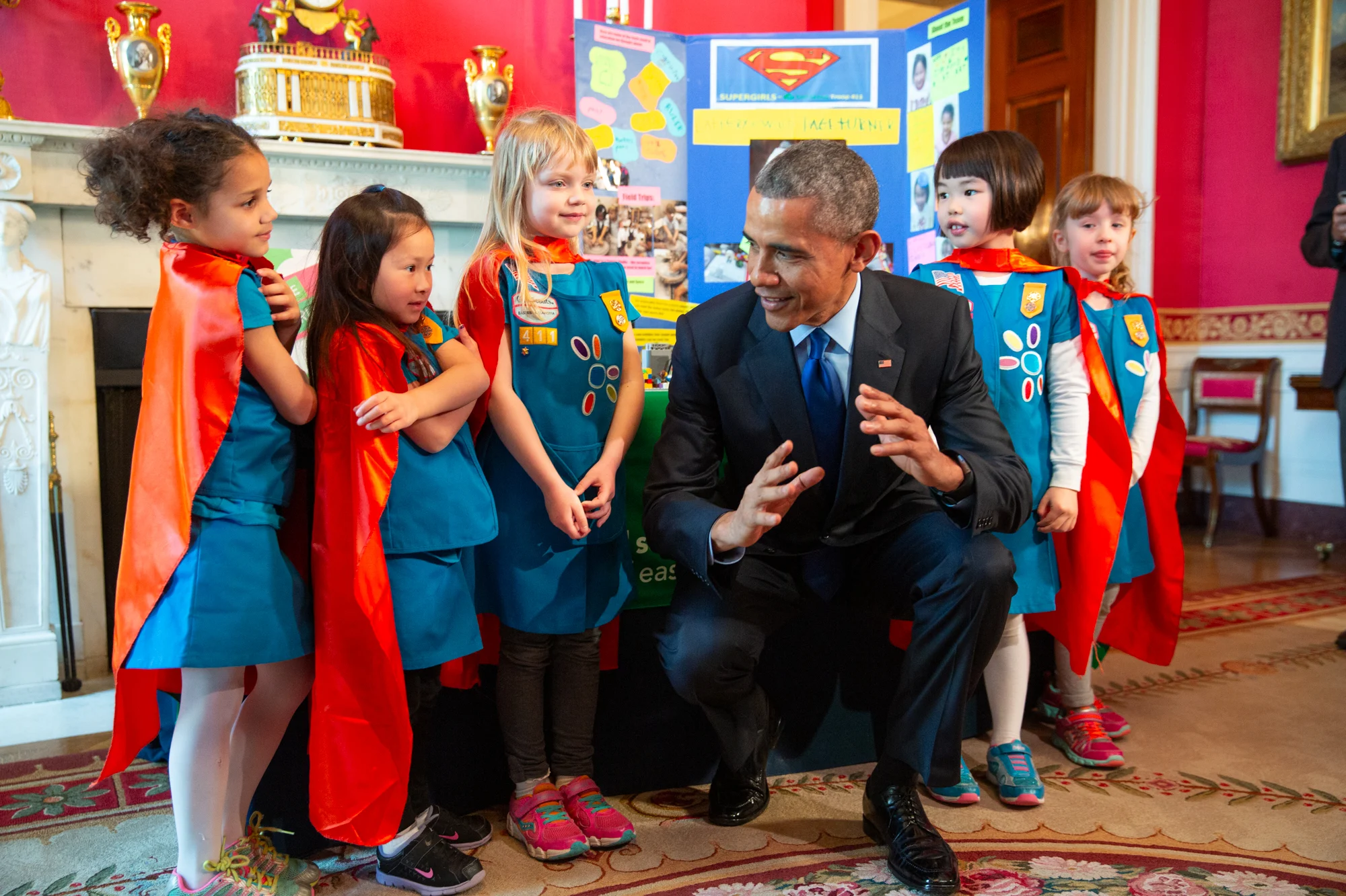 President Obama crouches down around five young people with a range of light and medium skin tones. All girls are dressed in green Girl Scout uniforms and red capes. 