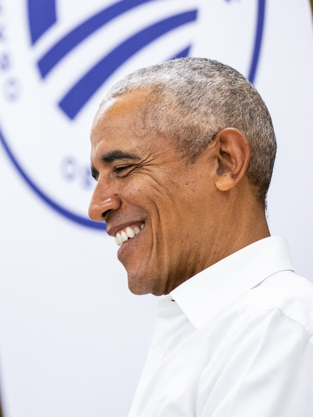 A side headshot of President Obama, smiling. He has a medium skin tone and gray hair. He is wearing a white button-down shirt.  Behind him is the Obama Foundation logo in blue.