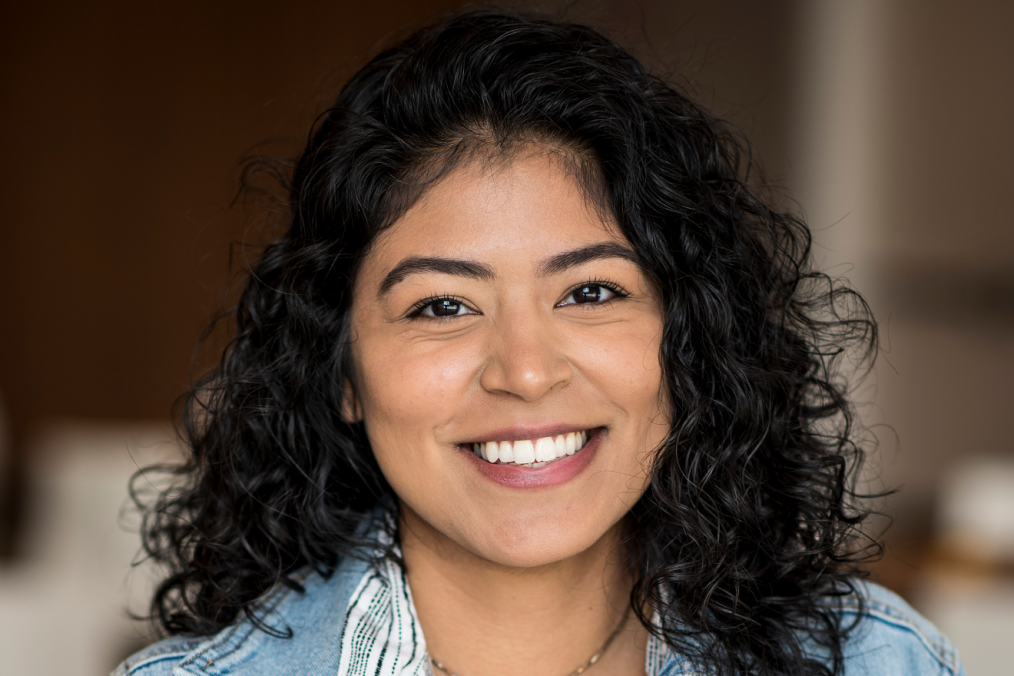 A headshot of a young woman smiling at the camera. The young woman has olive skin and brown eyes. She has dark curly hair that hits her shoulders and is wearing a blue denim shirt. 