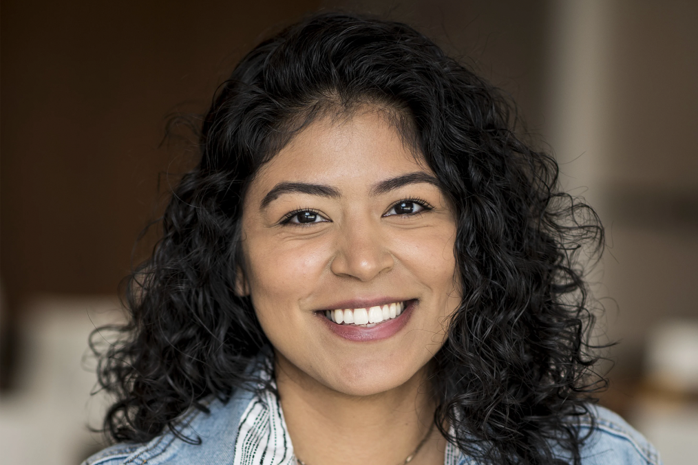 A headshot of a young woman smiling at the camera. The young woman has olive skin and brown eyes. She has dark curly hair that hits her shoulders and is wearing a blue denim shirt. 