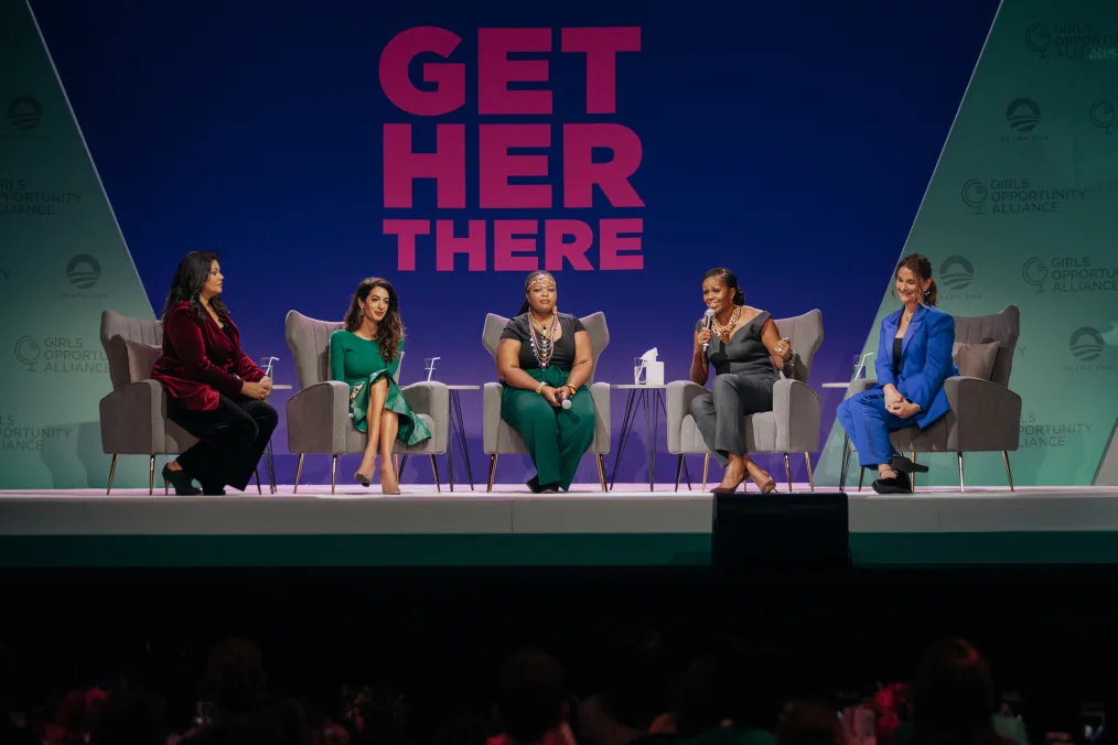 Sara Sidner sits on the left side of a stage listening as Michelle Obama speaks on a panel discussion seated next to Amal Clooney, Wanjiru Wahome, and Melinda French Gates. The speakers are a range of skin tones and wear dress clothes in an array of colors.The stage sign in the background reads, “Get Her There.”
