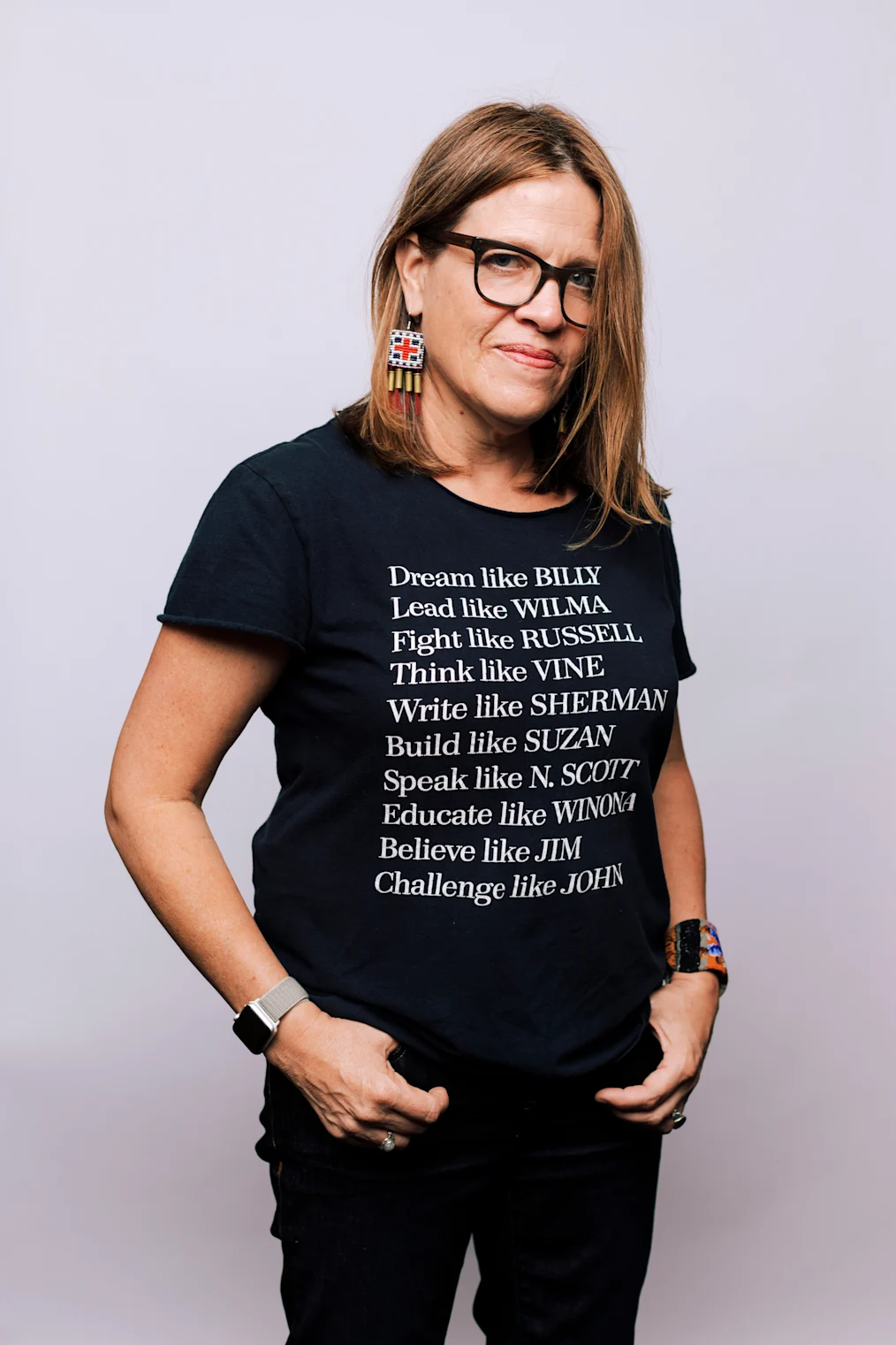 The image is a photographed portrait of Marie Watt. Marie is standing in a room with a white wall backdrop behind her. She is staring into the camera. She has a deep olive complexion and brownish red hair that falls to her shoulders. She is wearing black framed glasses and square earrings with a pattern inside them. The earrings also have tassels hanging off the ends of them. Marie is wearing a black tshirt that reads “Dream like Billy, Lead like Wilma, Fight like Russell, Think Like Vine, Write Like Sherman, Build Like Suzan, Speak Like N. Scott, Educate Like Winona, Believe Like Jim, Challenge Like Jon.” Each of the people referenced are Indigenous leaders, activists, and educators. She is wearing dark denim jeans.