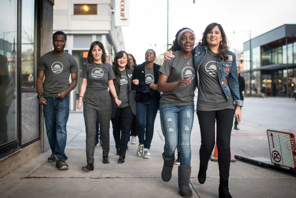 A groups of happy people wearing gray Obama logo t-shirts walk down a sidewalk in an urban area.