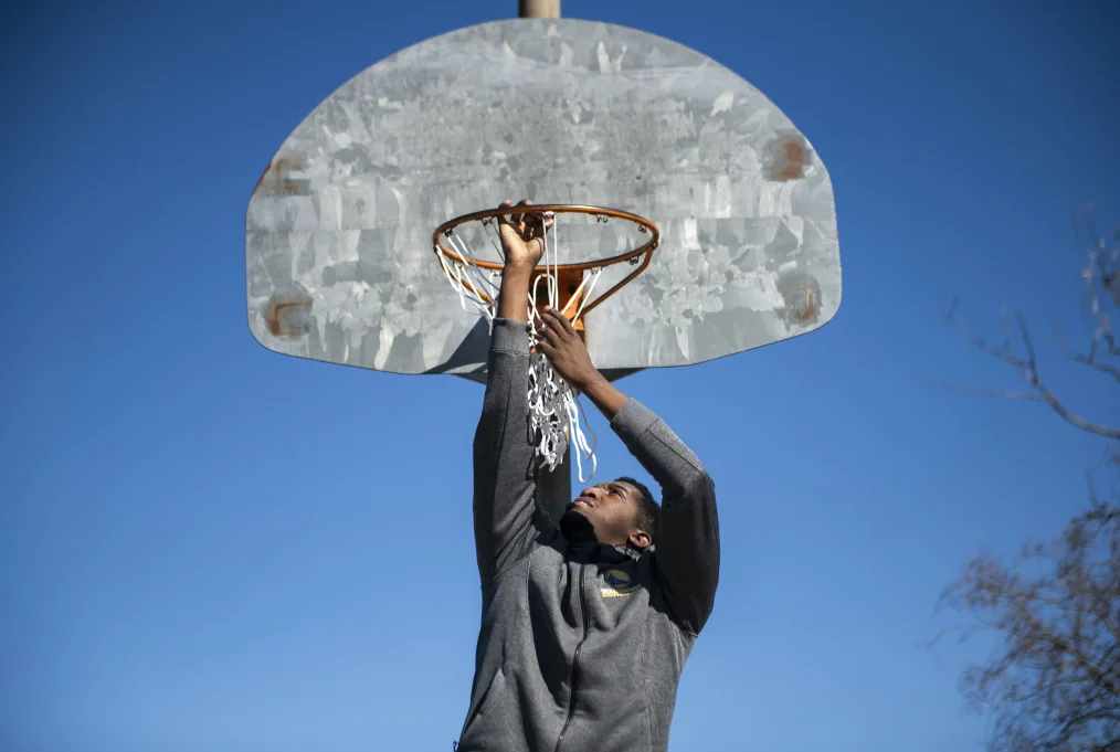 A young man with a deep skin tone hangs on the rim of a basketball hoop. He is wearing a gray hoodie.The blue sky is in the background.