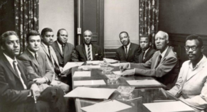 A picture of Martin Luther king jr and other men of color wearing suits sitting at a table in black and white with papers of the table in front of them 