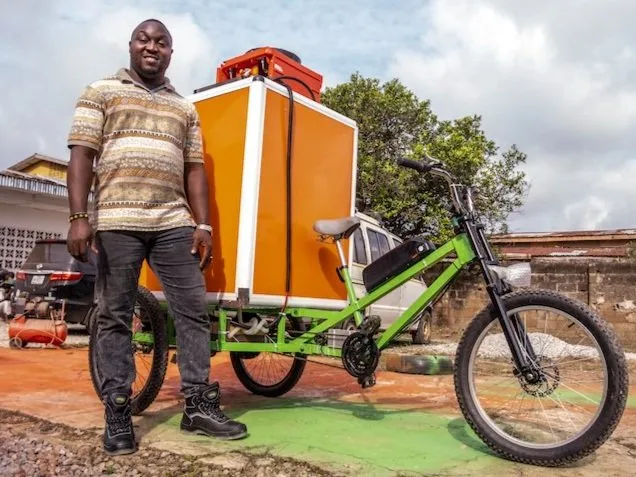 Tolulope Olukokun, a Black man with a dark skin tone and no hair, stands in front of an orange and white ThinkBikes. He is smiling. It is a sunny day. 