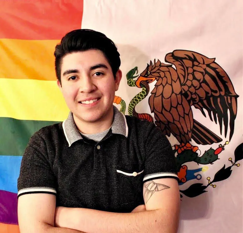 A male with a light skin tone takes a picture in front of a pride flag and the flag of Mexico