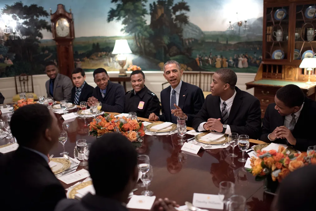 President Obama sits around a table with White House Mentorship participants. All young men are wearing suits and have a range of light to deep skin tones.