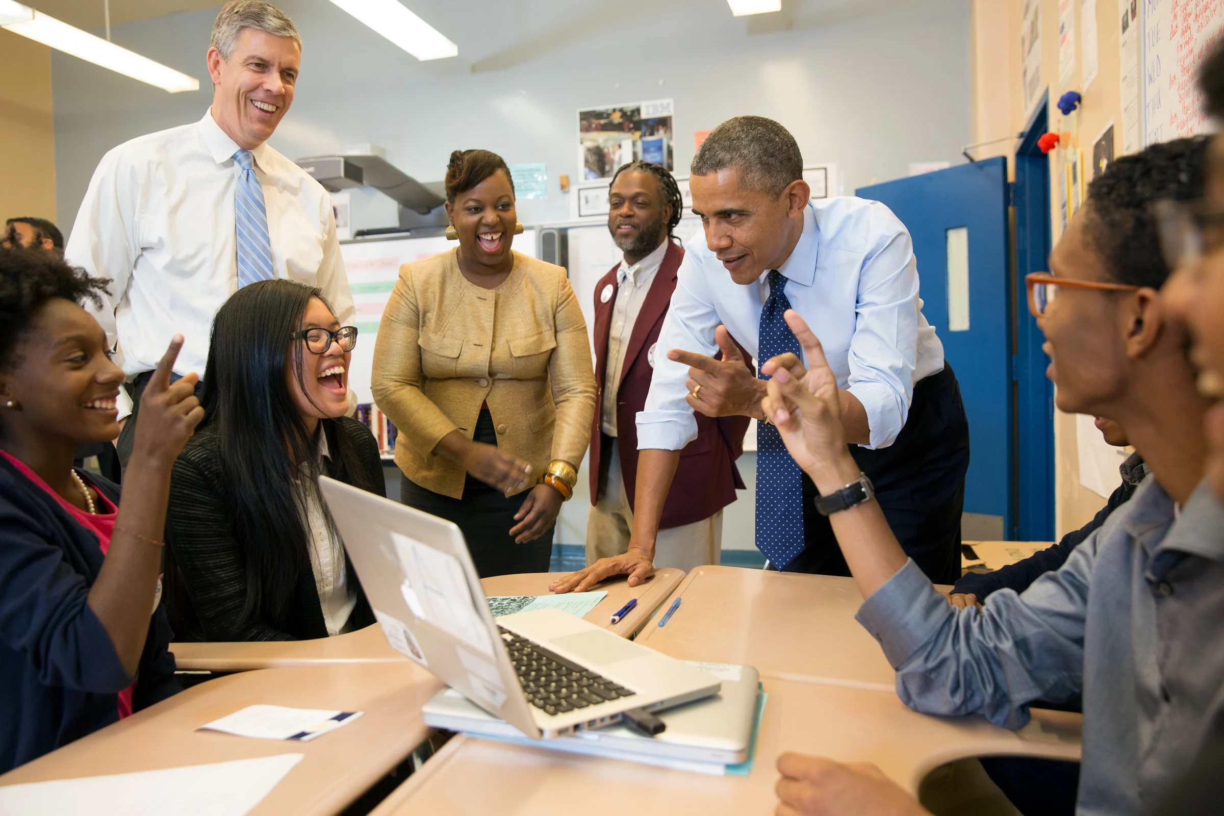 Barack Obama with one hand on a desk and the other pointing forwards looks at a group of people sitting and standing around him smiling and laughing. There is a computer on the desk. 