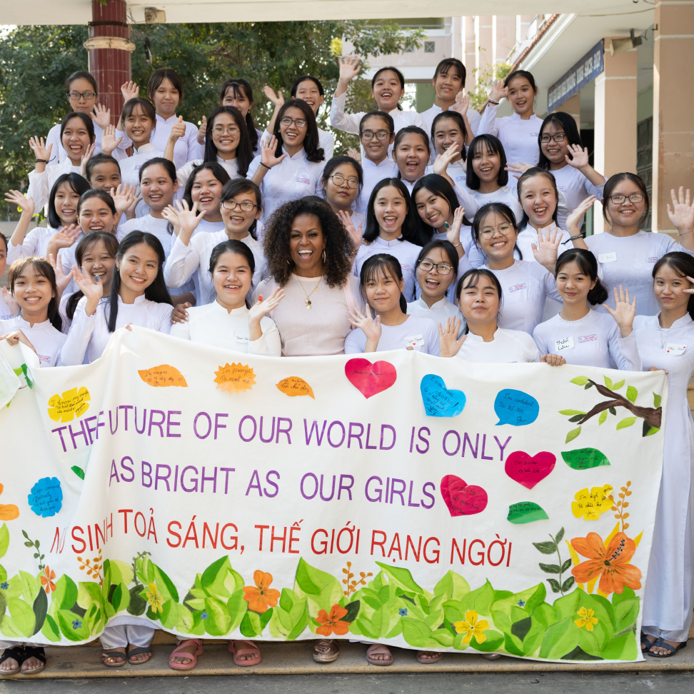 A group of young girls stand together posing for a picture. Michelle Obama stands in the middle of the group of young girls. The picture is outside. They are all holding up a large banner. The words on the banner are, "The Future of Our World is Only as Bright as our Girls" in English. Below the same message is written in Vietnamese.