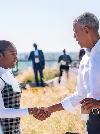 An image of Barack and Michelle Obama shaking the hands of two people while smiling. Barack Obama is shaking the hand of a teenage girl with a deep medium skin tone and dark hair pulled into a braid. Michelle Obama is shaking the hand of a woman with long braids. Barack Obama has a light deep skin tone and gray hair. He is wearing a light colored button down shirt with dark pants and holding a coffee cup in his left hand. Michelle Obama has a medium deep skin tone and curly dark hair. She is wearing an orange sweater and an orange shirt. The picture takes place outside. 